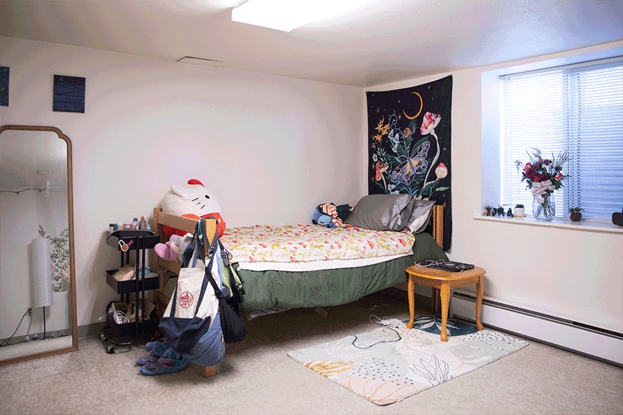 A cozy bedroom with a twin bed, small table, large window with blinds, a wall tapestry and other floral decor.